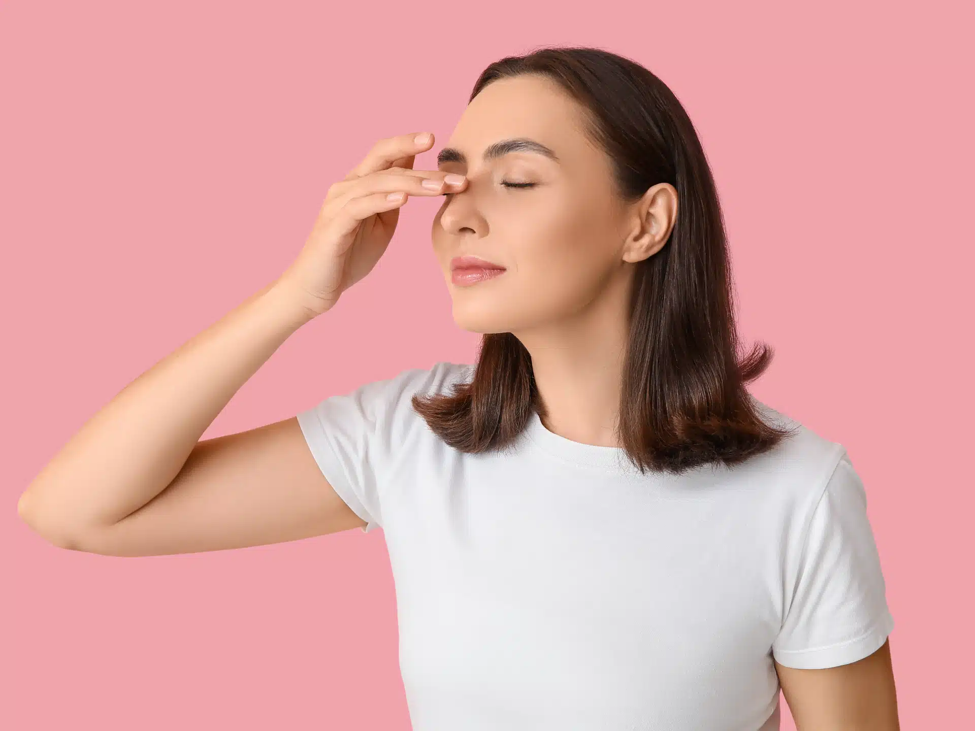 A woman with her eyes closed wearing a white t-shirt, gently touching the bridge of her nose with her fingers